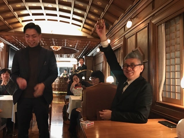 ななつ星 in 九州の車内で、手を挙げて笑うゲストと黒崎博斗（2017年）